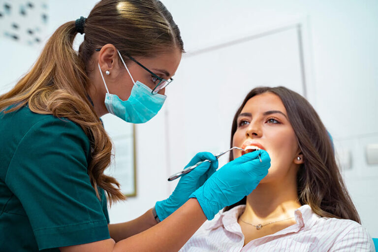 The image shows a dentist wearing a green uniform and blue gloves, attending to a female patient who is seated in a dental chair. The dentist is holding dental instruments and performing a dental examination or procedure. The setting is in a dental clinic.