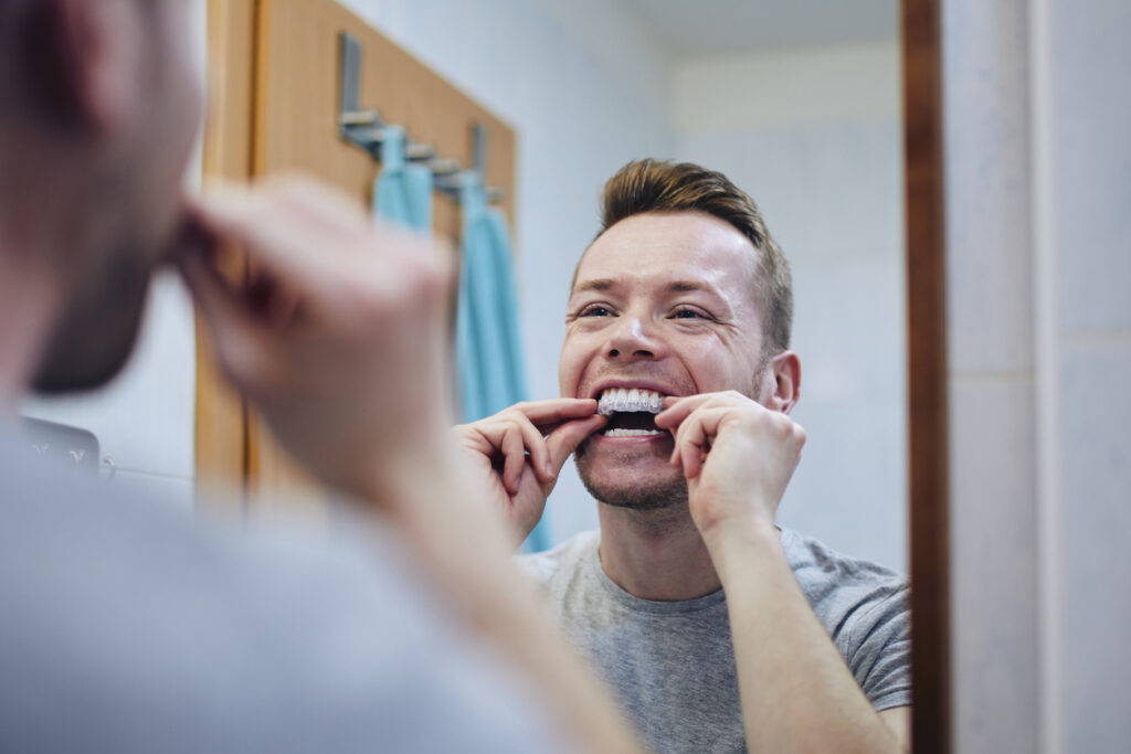 A young man is standing in front of a mirror wearing a gray shirt, holding a silicon tray with both hands. He is preparing to attach the tray to his upper teeth for a teeth whitening treatment. The scene emphasizes dental care and personal grooming, with the young man carefully positioning the tray.