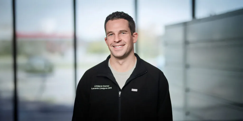 The image shows Dr. Laramie Lindgren standing while smiling. Dr. Lindgren is wearing a black jacket with a text on the left side of the chest. The background appears to be an indoor setting with large windows.